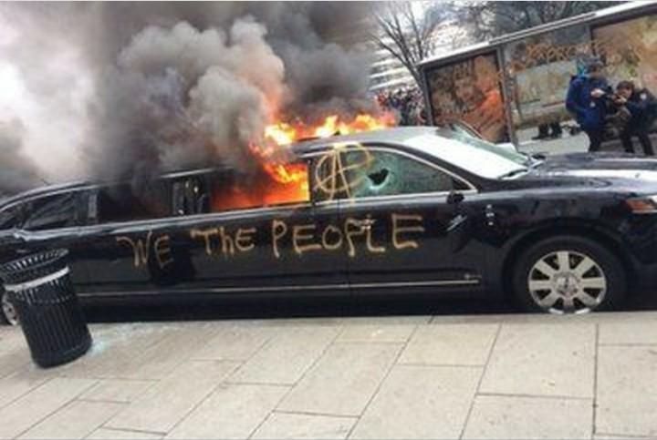Schwarze Stretchlimousine brennt, daneben ein schwarzer Metallmüllbehälter, über die Seite der Limousine steht: We the people
An der kaputten vorderen Scheibe ist ein A im Kreis.