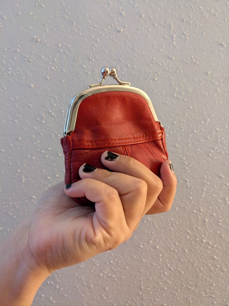 Photograph of a woman's hand with scuffed and worn black nail polish, gripping a dyed red leather coin purse with a metal clasp.