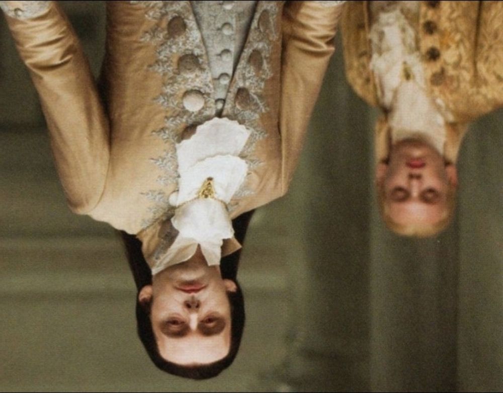 Aro aus der Twilight Saga in festlichem gold schimmerndem Gewand, der herablassend hinab blickt im Hintergrund eine weitere ebeso gewandete und blickende Personen  das Bild steht Kopf 