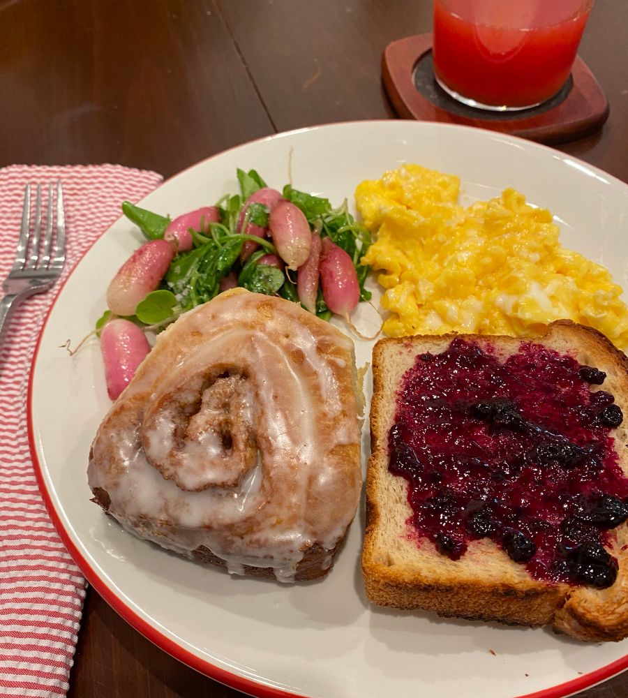 Breakfast plate with scrambled eggs, fried breakfast radishes with arugula, toast with blueberry jam, and a whole wheat cardamom cinnamon bun