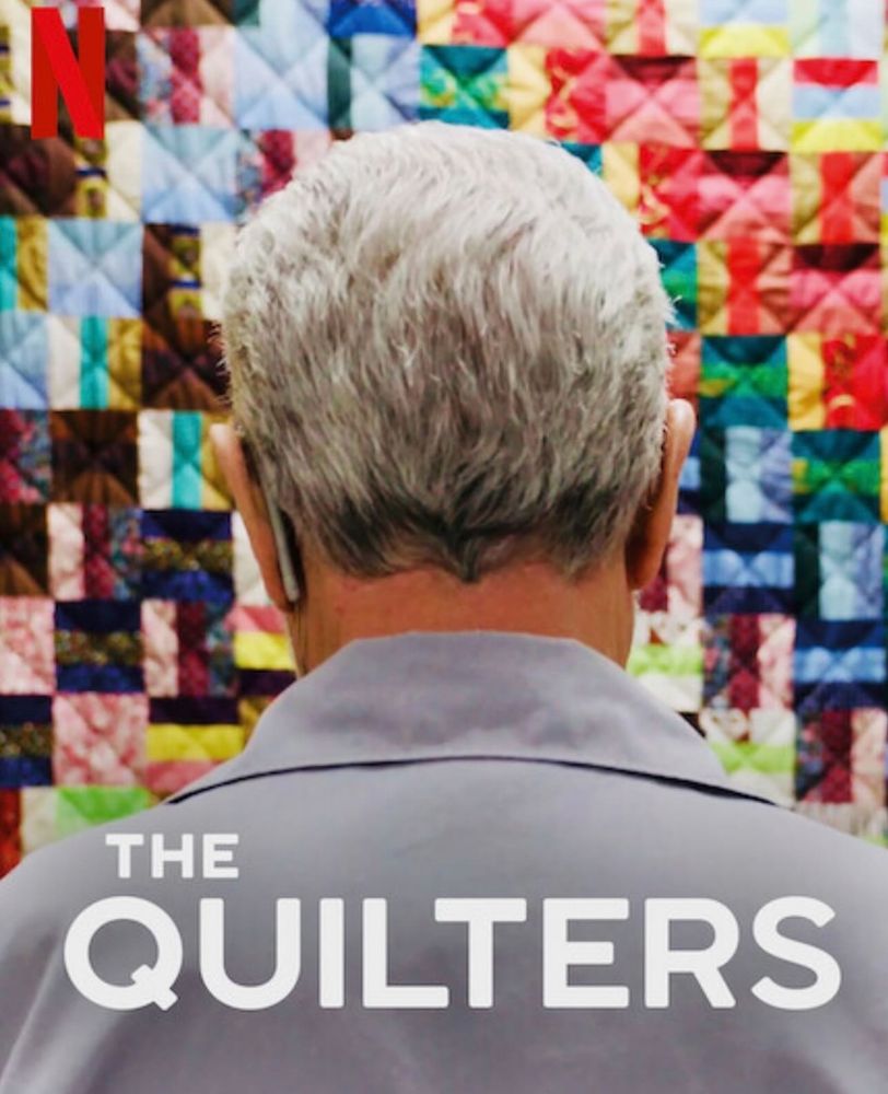 A white man with grey facing towards a multicoloured quilt. He shown from the back of the head. Text reads 

THE QUILTERS
The letter N is in read in the left hand corner. 