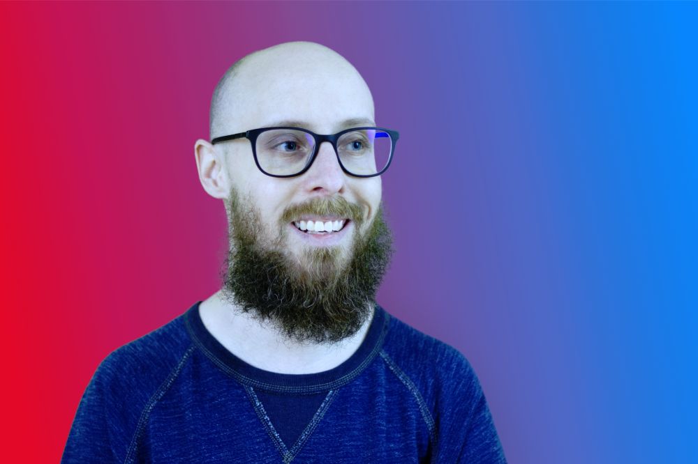 Tristen Grant smiling in front of a red-to-blue gradient background. He has white skin, a bald head, glasses, and a full beard, and is wearing a dark blue shirt with contrast stitching.
