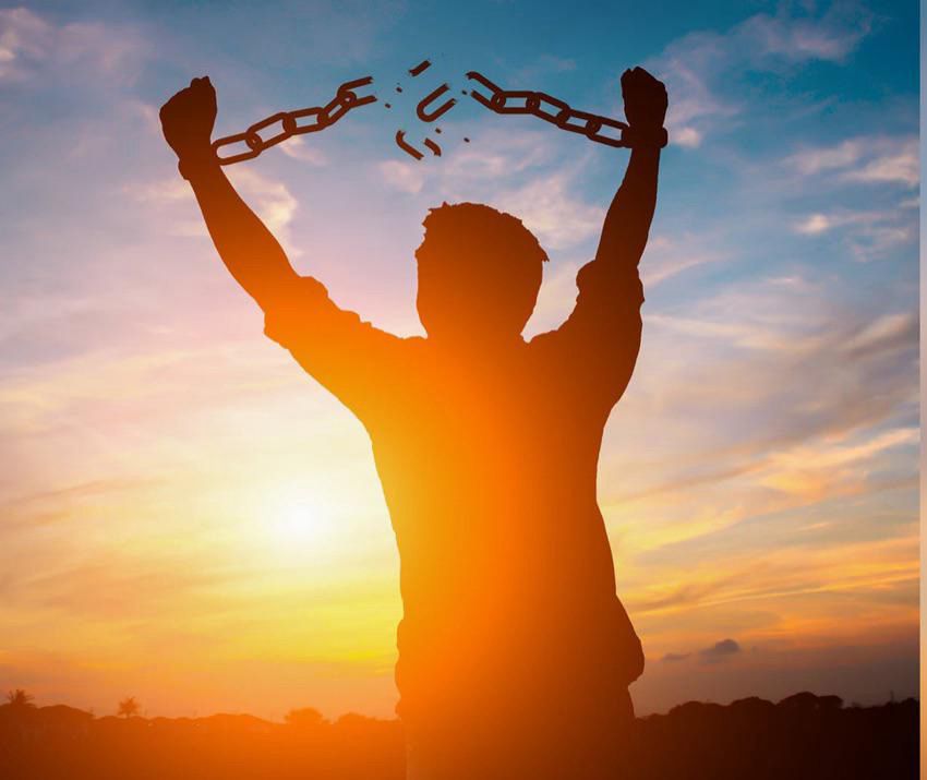 Image of a man’s silhouette who has his arms up in the air who is chained, but the chain has been shattered in the middle. There is a sunset and the silhouette of a horizon it’s peaking up from.