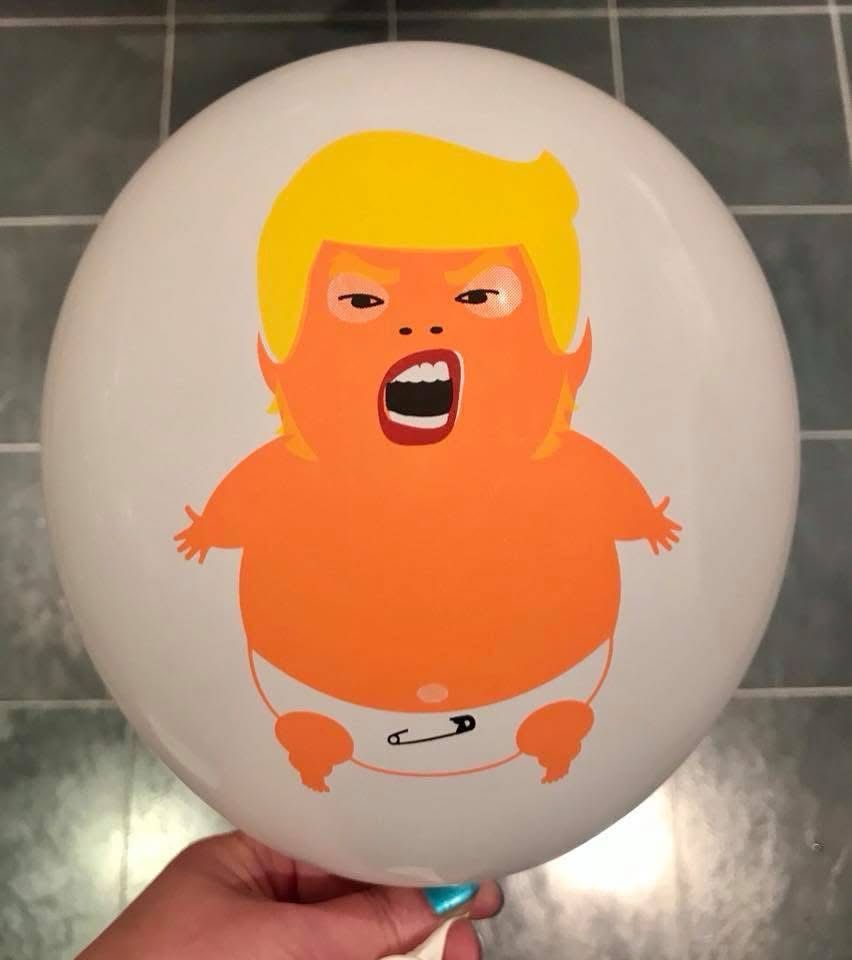 A baby trump mini balloon, inflated and held for a pic in my kitchen. The balloon is white with an orange baby trump in a white nappy in it, he has tiny hands and an open mouth probably spouting nonsense.