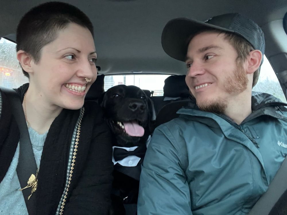 Lilia has a buzzed head, black soft jacket with two lines of diamond shaped embroidery in cream and blue and a light blue knit sweater on under it. They have a septum piercing and a gold moth or butterfly necklace. She’s looking from the left at Chris sitting on the right and both are wearing a seatbelt sitting in a vehicle. Chris is wearing a blueish-grey-green rain jacket and grey trucker hat and has a medium light beard. Both are smiling big at each other. Their dog Nova, a black lab is wearing a collar with a shirt front and black tie. His tongue is sticking out and he looks excited.