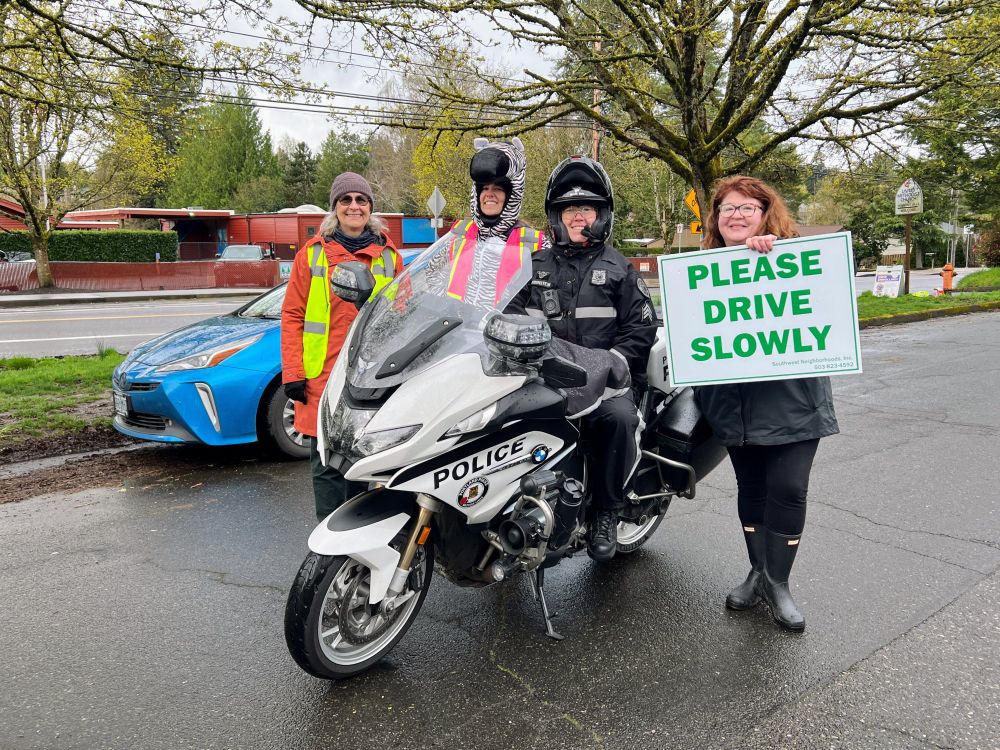 A woman holds a sign Please Drive Slowly and stands with two others with a Portland Police motorcycle officer