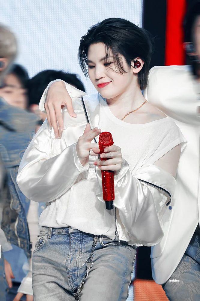 Lee Jihoon wearing a silk white jacket over a tank top holding a red mic with someone off camera’s arm around him looking incredibly girlfriend.