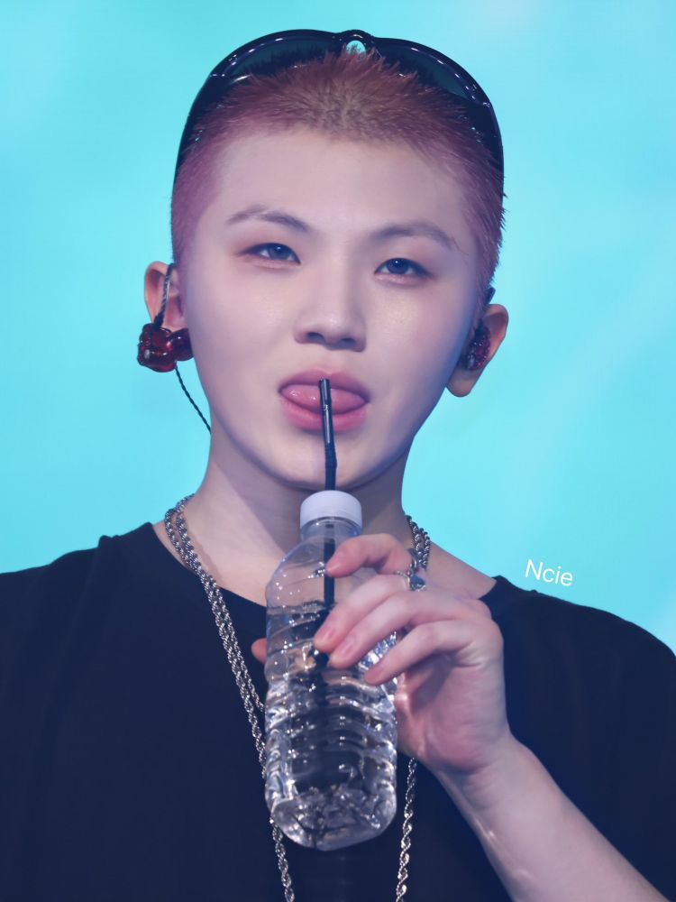 Lee Jihoon with a shaved head looking perfectly round and sticking his tongue out against the straw of his water bottle. 