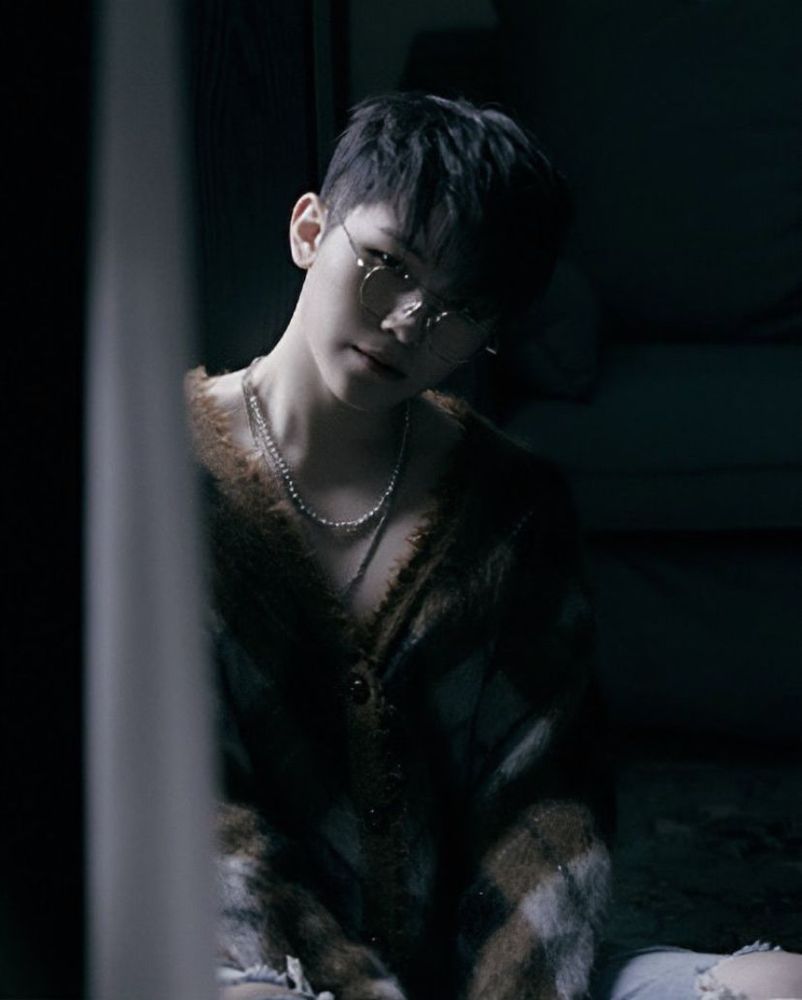 Lee Jihoon wearing a v neck plaid sweater and glasses, harshly shadowed while looking at the camera.