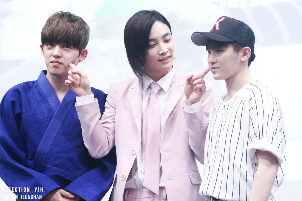 Yoon Jeonghan wearing a pink suit, smiling and reaching out both pointer fingers to poke Choi Seungcheol and Lee Jihoon's cheeks. They both look incredibly unamused.
