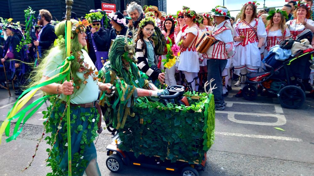 A woman with a staff accompanies a seated mask figure in a foliage-enhanced mobility scooter of some kind