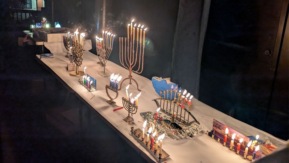 A table full of menorahs of all different shapes and sizes. 6 candles are lit on each. 
