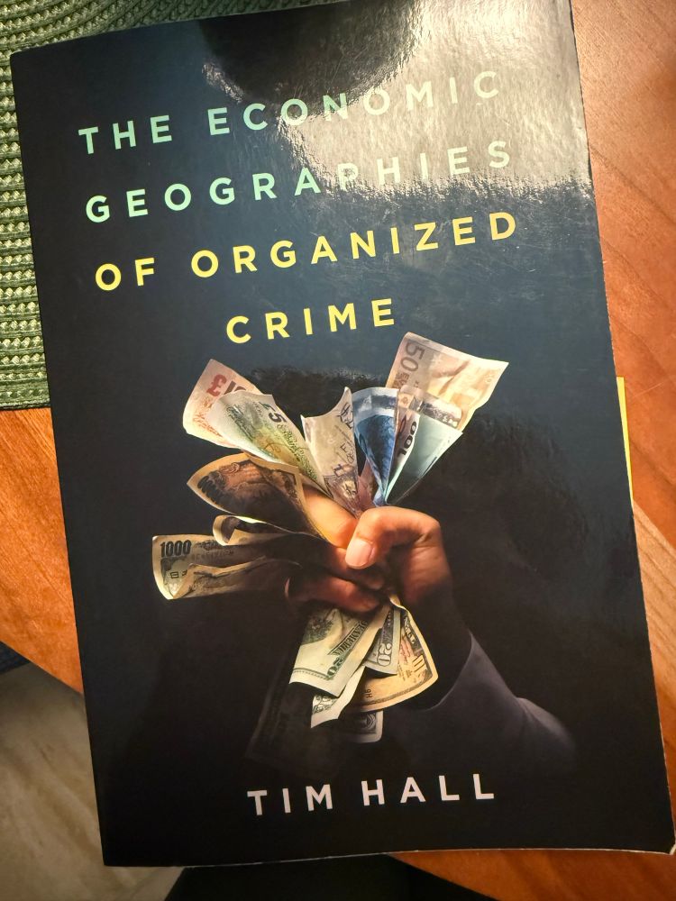 Book title: the economic geography of organized crime by Tim hall with a cover picture of someone’s hand gripping cash in different currencies 