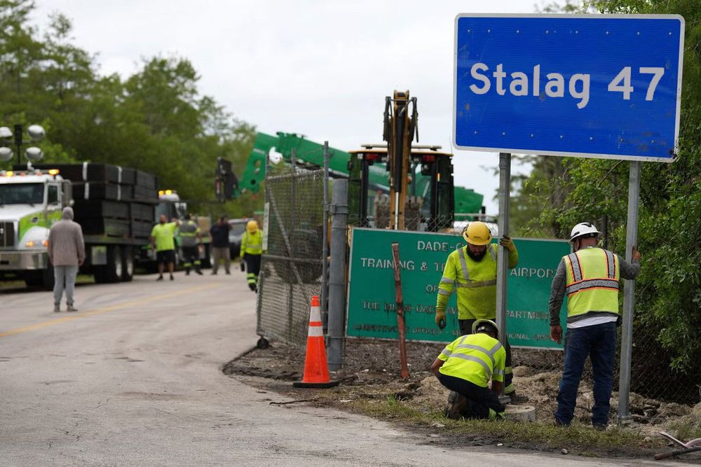 Florida highway workers erecting a blue “Alligator Alcatraz” sign, which I photoshopped into “Stalag 47