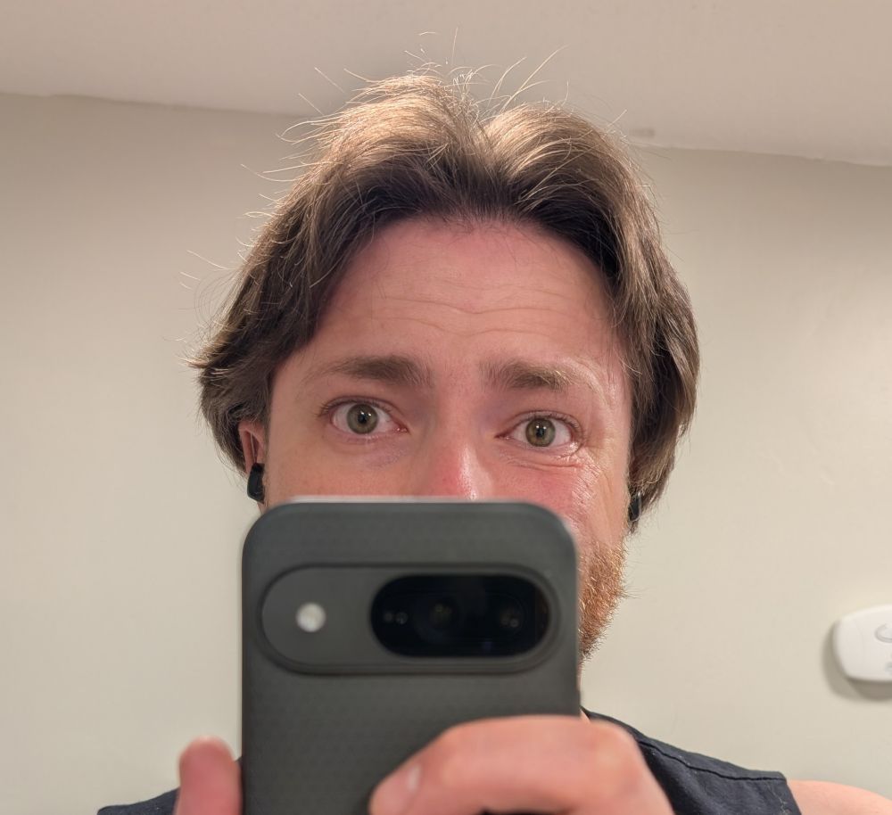 An up close mirror selfie of a man with the phone covering his face below the bridge of his nose. His hair on the left flairs outward in little wisps, while his hair on the right is lying flatter against good head.
