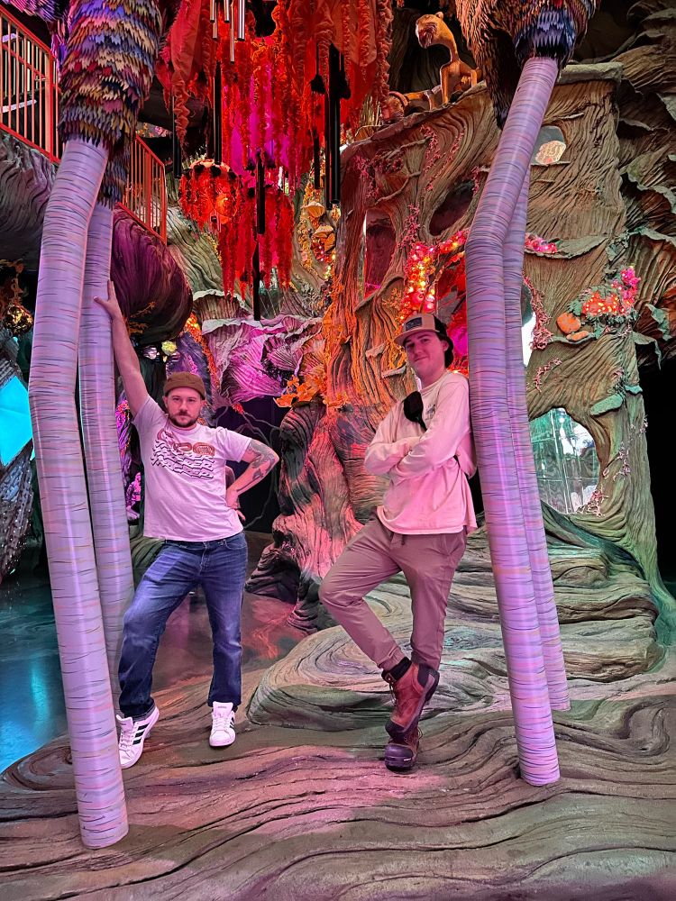 Two men standing under a colorful creature in a weird forest-esque area