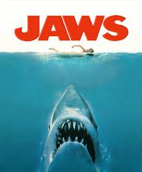 Poster for the movie Jaws - there is a large shark at the bottom of the poster seemingly swimming up to a woman swimming on the surface of the ocean