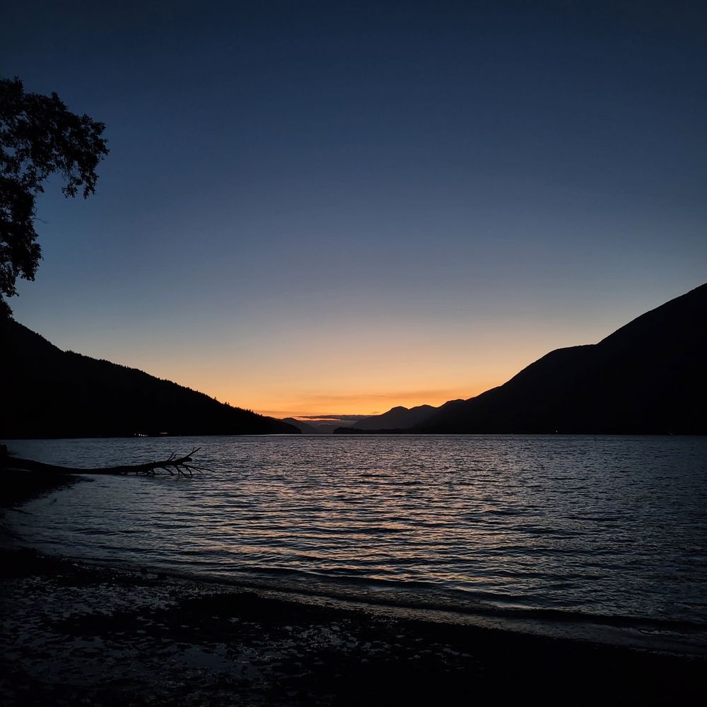 The last warmth of sunset creeps down behind the silhouettes of mountains in the background. In the foreground, the faint light reflects upon a lake and its shore.