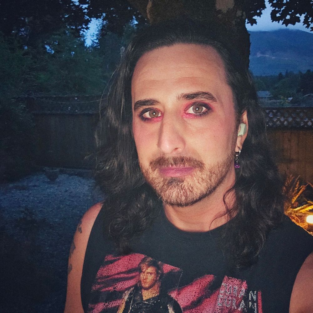 An overprocessed selfie (high brightness, low contrast, and grain). My hair growth is noticeable, just past my shoulders in some sections. I'm wearing a mix of black and red eyeshadow, with a touch of red sparkles at my inner eye area. I'm also wearing a small amount of mascara, and have a purple dangling bead earring in my left ear. I'm listening to music with my mint-coloured earbuds, and smiling slightly with my mouth closed. My sleeveless Duran Duran band tee is just noticeable, the photo cutting it off at my chest. In the background, just beyond a backyard wooden fence, you can make out some trees, a mountain, and the cloudy skies above.