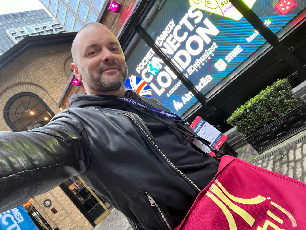 Stephen Reid, VP of Learning at The Endless Foundation stands in front of an illuminated sign that reads Pocket Gamer Connects, London. He is wearing a black hoodie, black leather jacket and  red Atari branded bag. 