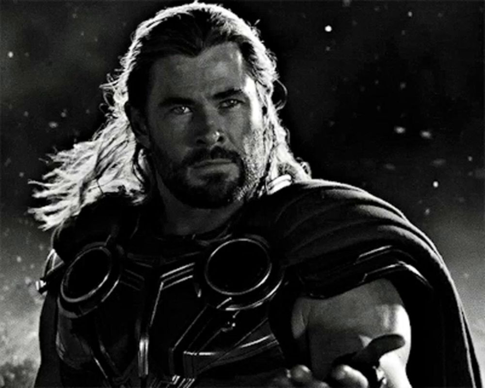 a black and white photo of a man with long hair and a beard wearing a cape and goggles .