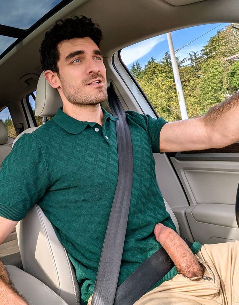 A man dressed in a green polo and khaki pants is driving with one hand on the wheel. His thick cock is fully erect and protruding from the fly of his pants.