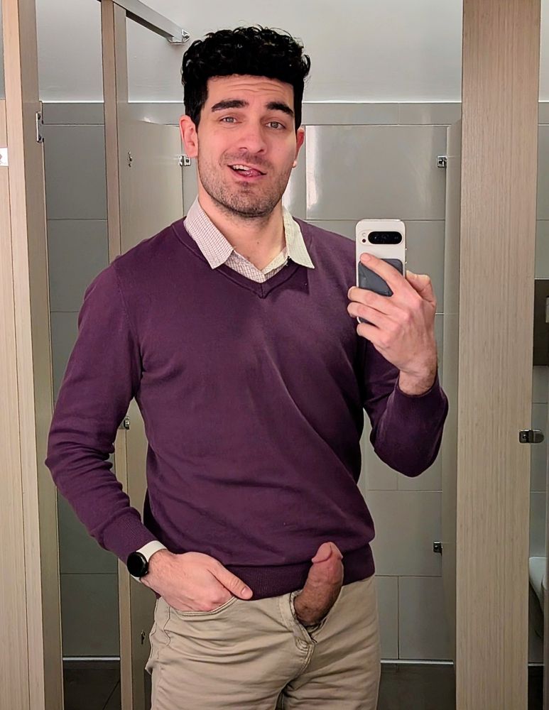 Office restroom mirror selfie. A man in a purple sweater vest and khaki pants stands with a knowing smirk. His thick, protruding cock extends from the open fly, curving upward with evident rigidity.