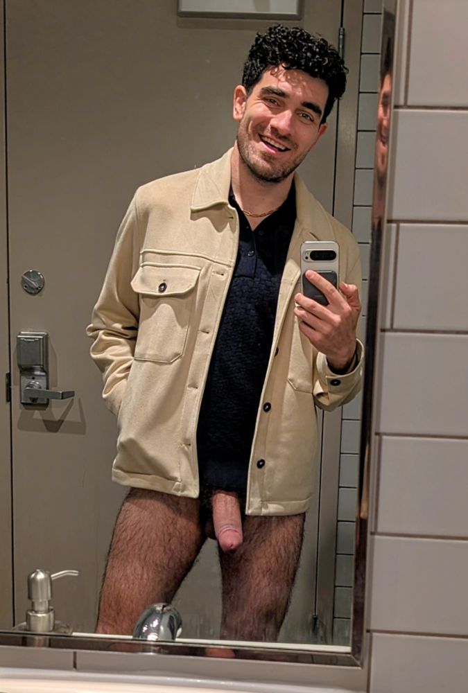 A bathroom mirror selfie of a smiling man with short curly hair. He's wearing a dark blue polo, cream jacket and nothing else. His thick engorged cock and hairy legs are  fully exposed and on display.