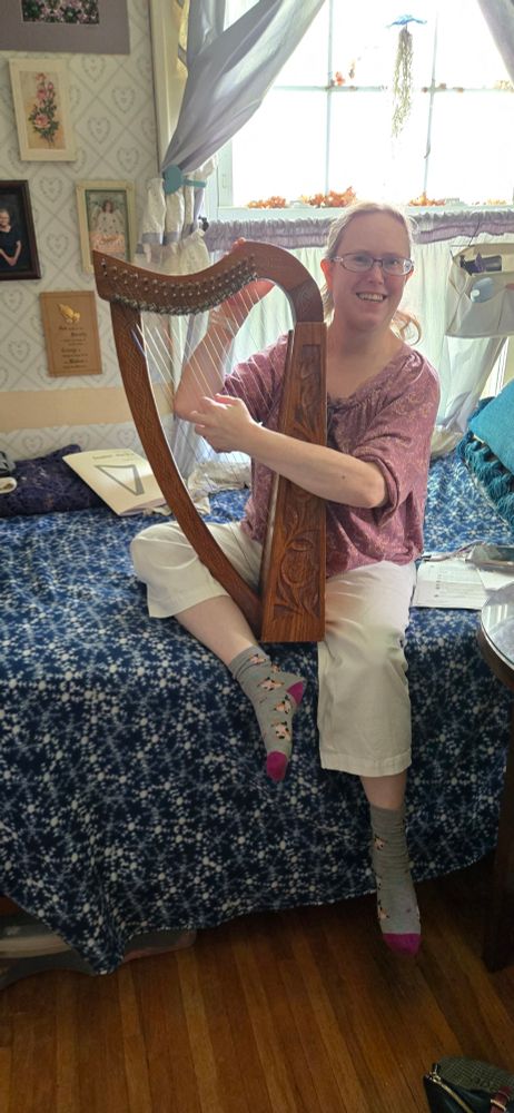 A woman sitting on a bed. One leg is down over the side and the other is pulled up next to the other knee. A large folk, or lever, harp sits between her legs. She holds it with one hand while the other is across the strings. 
