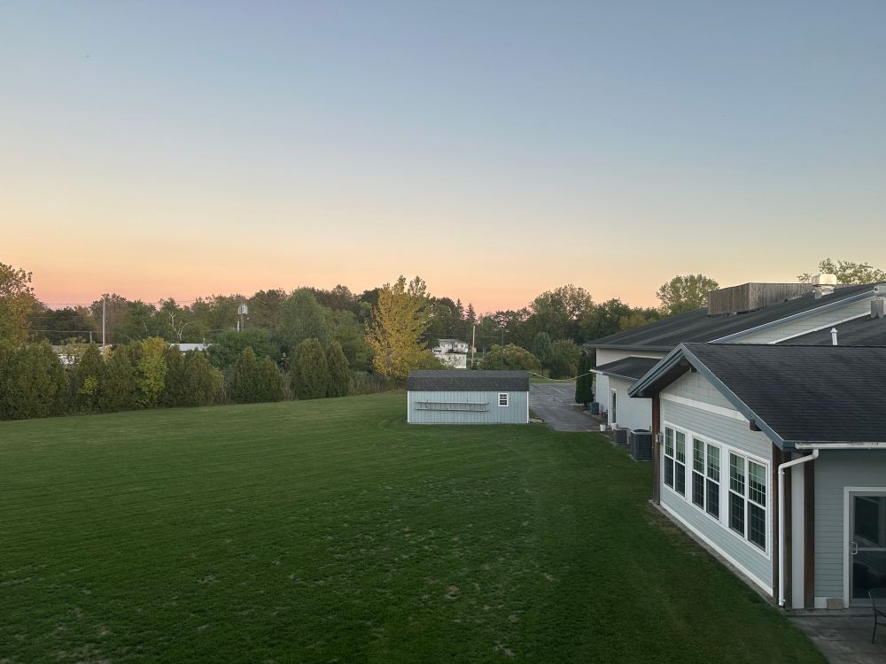 A green grass lawn with a grey building to the right and a shed in the middle distance. Distant trees are starting to show fall color and the sky is in sunset colors. 