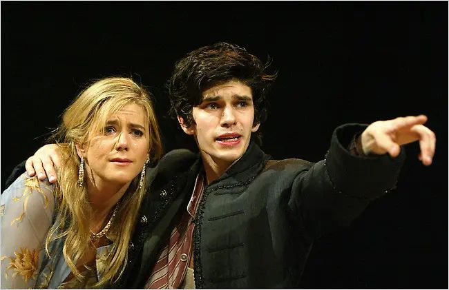 A photograph of a play in performance. Head and shoulders, on the right a young man with dark hair wearing a black jacket, points with his left hand forward and out of frame; he looks distressed. He has his right arm around a woman with long blonde hair; she is looking where he is pointing as if she can't see anything.