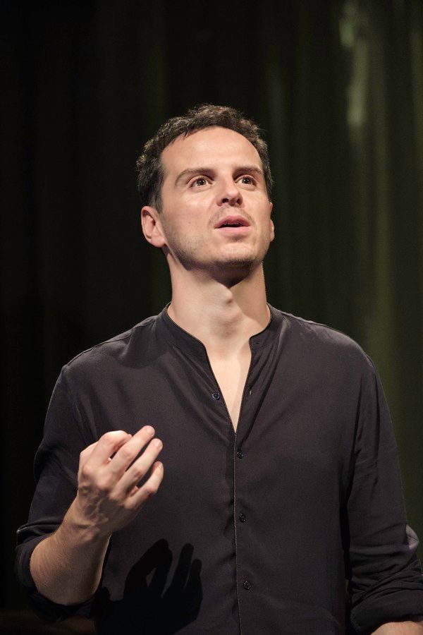 A photograph of a character in a play. Half length, a young man with short dark hair and dark eyes speaks, looking up slightly; his right hand is raised in front of him .He is wearing a plain black shirt with the sleeves pushed up and a couple of buttons undone.