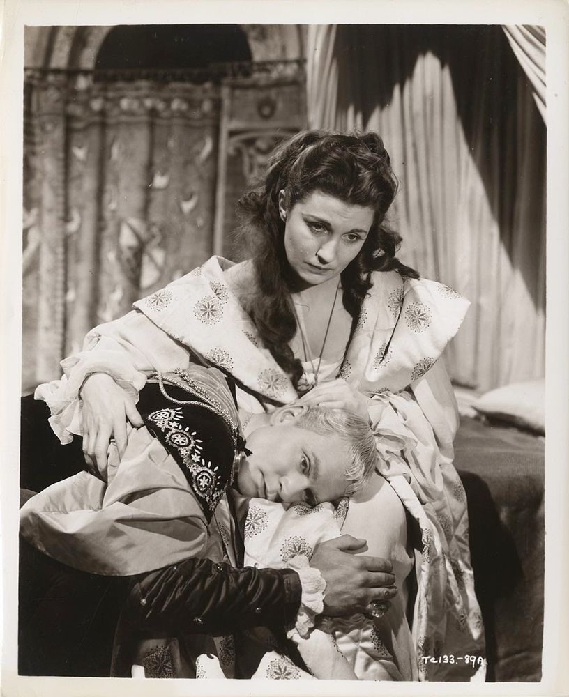 A black and white still from a film. A young man wearing an elaborately embroidered doublet is lying with his head in the lap of an older woman, wearing an elaborate satin dressing gown. Her hair is partly loose; he has his arm around her knees, she has her arm around his shoulders. They both look exhausted.