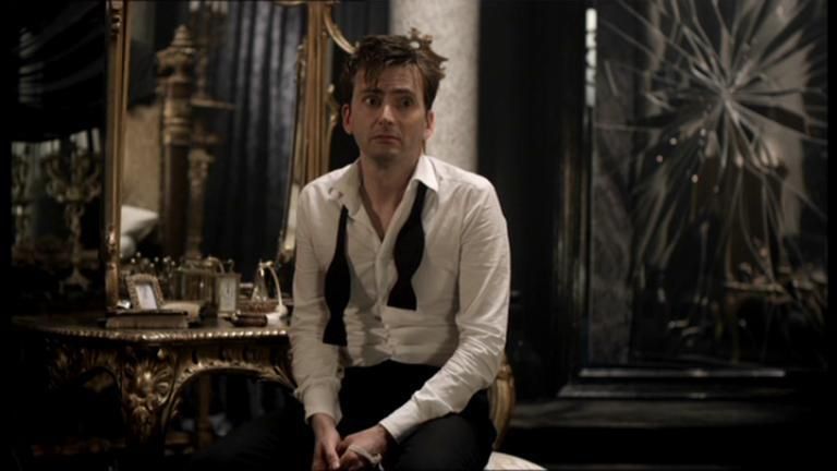A still from a film. A young man looking dishevelled, wearing a white shirt with the collar open and an undone black tie, is sitting in front of an ornate gilded dressing table with a mirror. There is a smashed full length mirror in the background. He looks stunned.