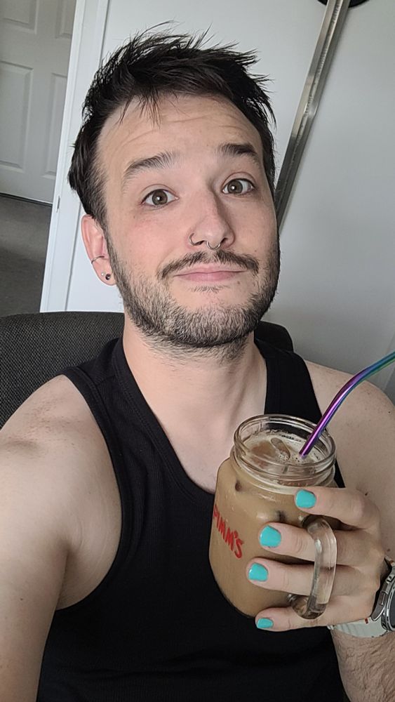 Picture of a man sat in a computer chair, taking a selfie, holding a glass of iced coffee.