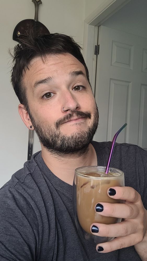 Picture of a man with messy hair wearing a grey t shirt, looking at the camera with a resigned expression at the fact that it is, once again, monday. He is also clutching an iced coffee.