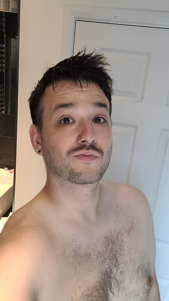 A topless man stood in a bathroom, looking at the camera,  having just shaved the beard back to stubble