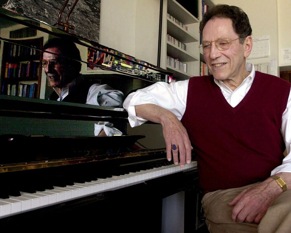 An elderly man with glasses and a burgundy sweater vest sits at a grand piano, smiling. His reflection is visible in the glossy black lid of the piano, showing him mid-laugh. Shelves filled with books and CDs line the background, suggesting a cozy and intellectual home setting.