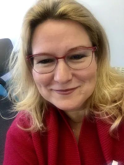 "A picture of Michelle Post, LMFT, smiling and with long blonde hair wearing red glasses and a red sweater, seated indoors against a light background."