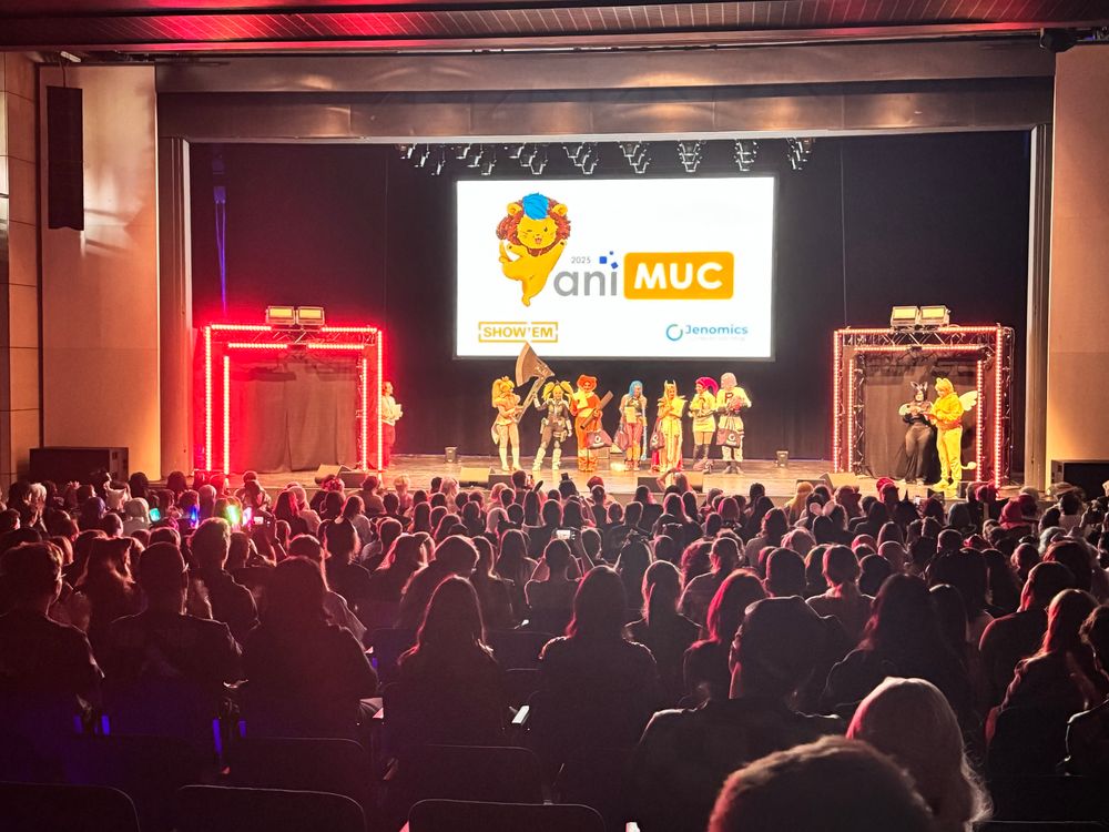 A crowded auditorium features performers on stage dressed in colorful costumes, with a large screen displaying the event logo for "aniMUC 2025." The audience, engaged and clapping, is illuminated by stage lights and colorful glow accessories.