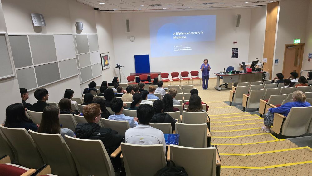 A female doctor in purple scrubs gives a talk to a lecture theatre full of young people who are aspiring to a career in medicine.