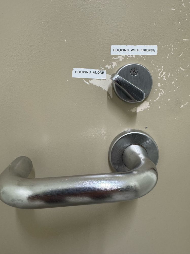 The chrome door handle on the inside of a bathroom door. The lock mechanism is unclear. So someone has added stickers saying pooping alone and pooping with friends. 