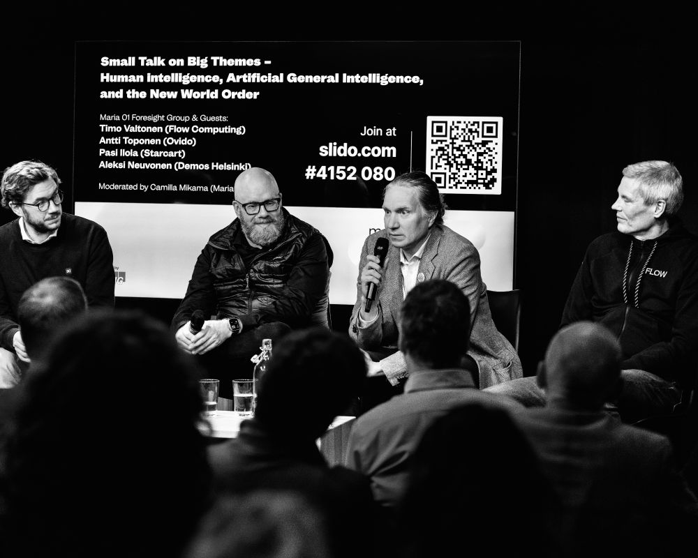 Black-and-white photo of the Maria 01 NEXT panel “Small Talk on Big Themes – Human Intelligence, Artificial General Intelligence, and the New World Order.” From left to right: Antti Toponen (Ovido), Pasi Ilola (Starcart, speaking into a microphone), Aleksi Neuvonen (Demos Helsinki), and Timo Valtonen (Flow Computing). Audience members are visible in the foreground, facing the panelists.