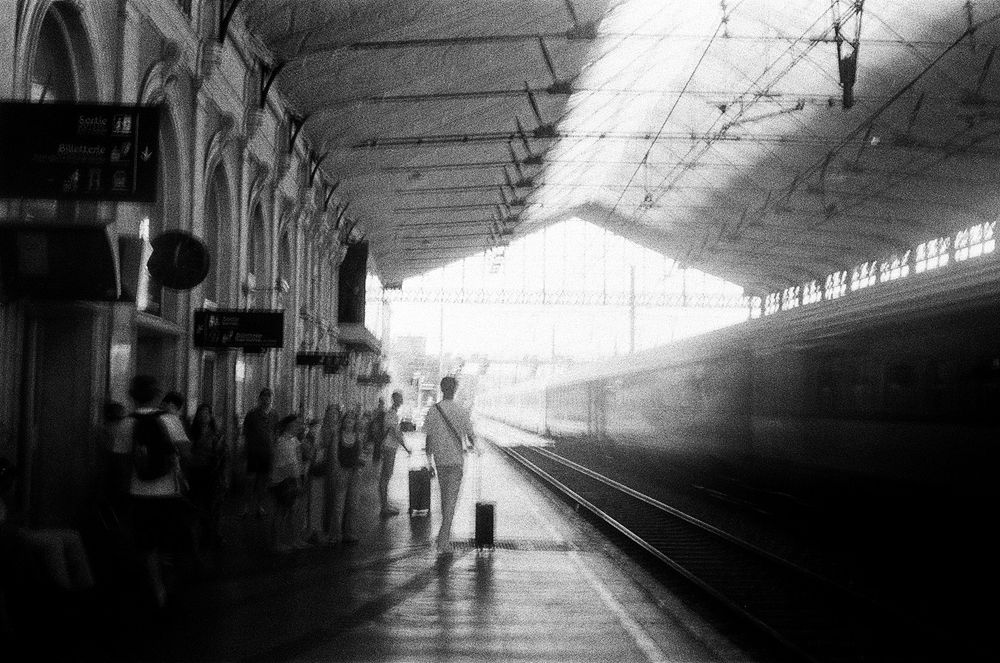A French train station. A man walk on the platform to the day light straight ahead with his luggage. A train on the right on another rail. The light reflects on the ground. Entrance to the hall is on the left. It's in Narbonne, south of France. 