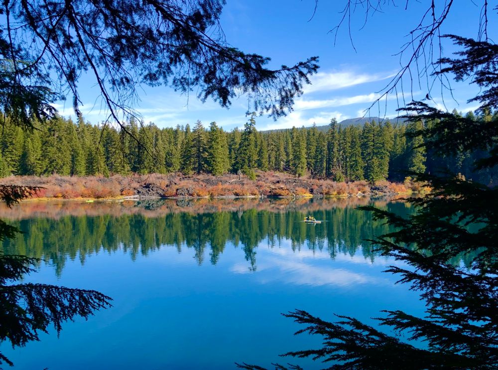 Clear lake's water is remarkably clear, allowing visibility to the bottom (over 100 feet in some spots). large pine trees and fall foliage are reflected in the lake’s water. blue sky overhead. 