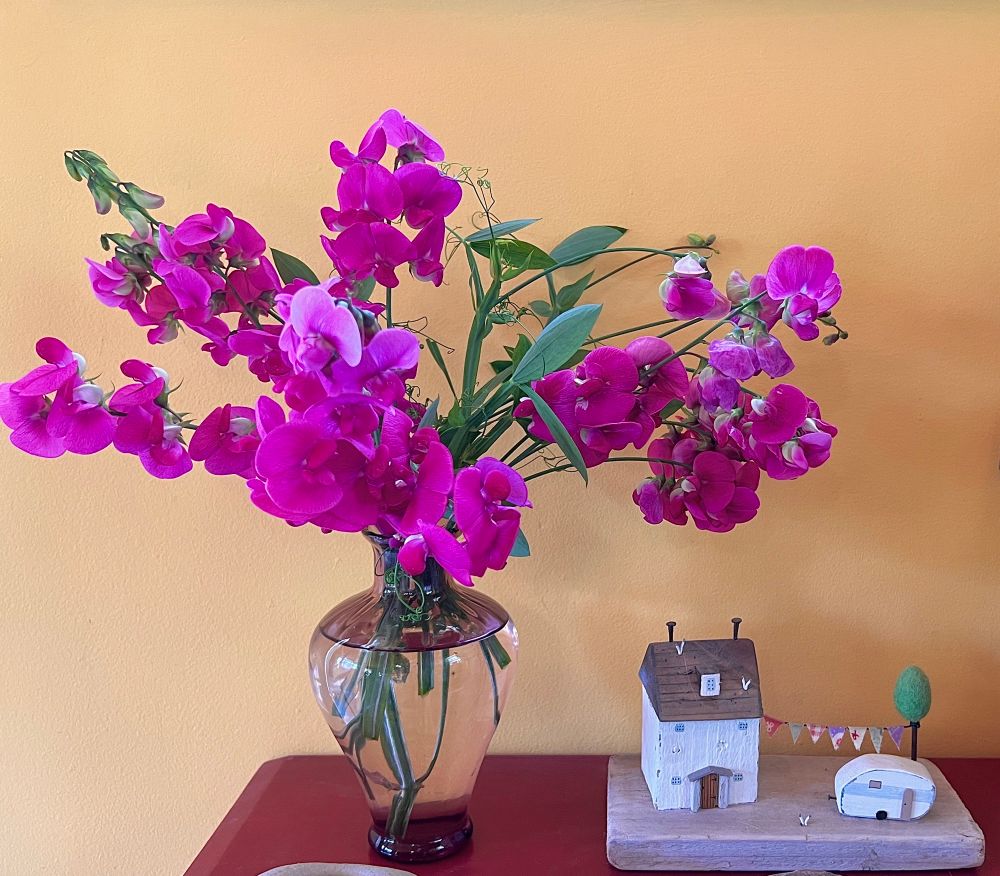 sweet peas in a vase on a red table. next to the vase is a little wooden display of a beach cottage with a camper, a tree and a cloth line. 