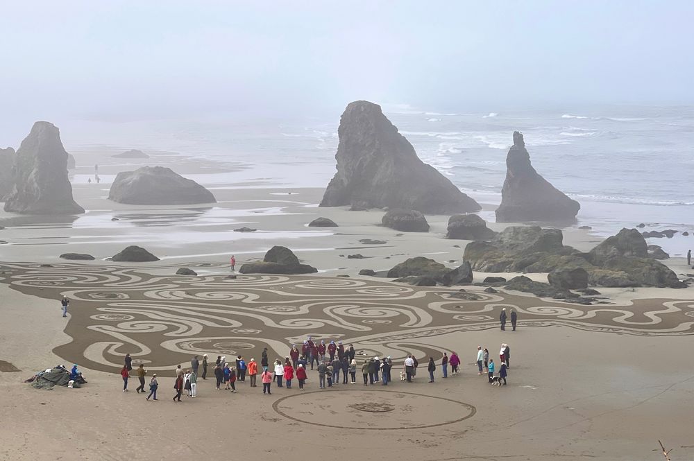 a large group of people on the beach ready to walk the labyrinth of “Circles in the Sand”, which is held every Summer in Bandon, Oregon. numerous large and small sea stacks in the background and a foggy sky above. 
