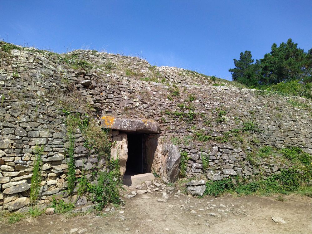 The entrance to ancient Gavrinis tomb, a stone wall built up around a dolmen entrance to a human-built cave. In front, the ground is dry, devoid of vegetation, overhead the sky is clear of clouds. Another hot day to come...