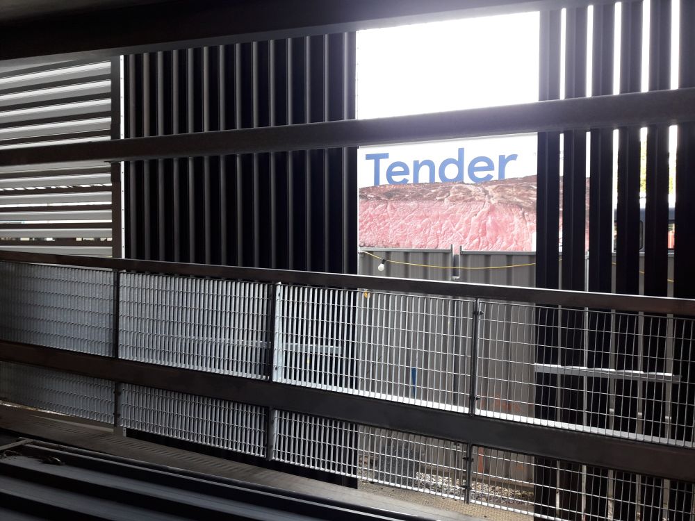 Through a gap in a range of steel grilles there is a billboard showing a cooked cut steak and the word tender
