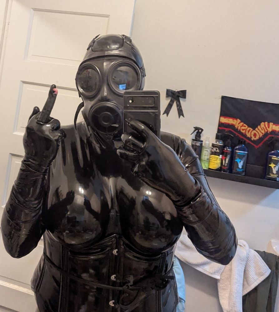 Nyxie giving the finger in a torn latex glove in a full drone outfit